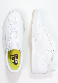 Ett par vita Reebok-sneakers med vita snören, perforerade detaljer och gula innersulor märkta "Reebok FACE Stockholm."