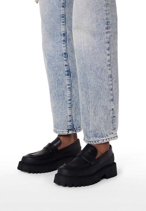 Schwarze Plateau-Slipper mit glattem Lederfinish und klobiger Gummisohle, kombiniert mit hellblauen Jeanshosen.