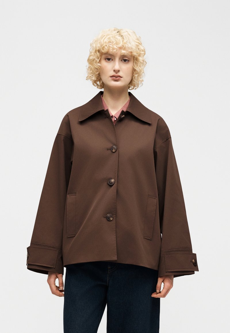 Γυναίκα με σγουρά ξανθά μαλλιά φοράει σκούρο καφέ σακάκι με κουμπιά, φαρδύ γιακά και μεγάλες μανσέτες, συνδυασμένο με σκούρο μπλε παντελόνι.