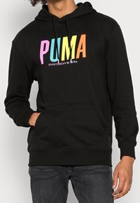 Persona che indossa un hoodie nero con il testo colorato "PUMA" e tasca frontale, abbinato a jeans scuri, con una mano parzialmente in tasca.