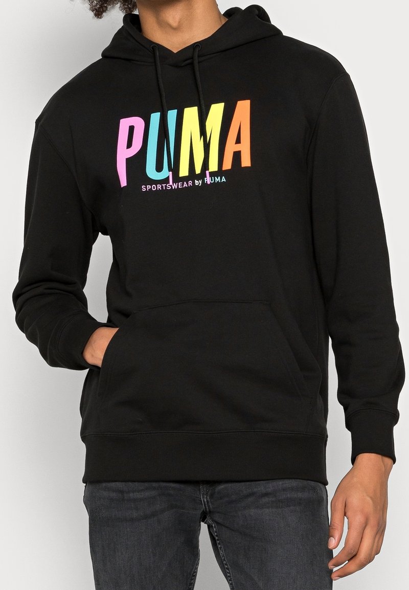 Persona che indossa un hoodie nero con il testo colorato "PUMA" e tasca frontale, abbinato a jeans scuri, con una mano parzialmente in tasca.