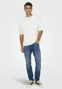 Suéter blanco de punto y manga corta con escote en V, combinado con jeans azules y zapatillas marrones con detalles en blanco. Diseño simple y casual.