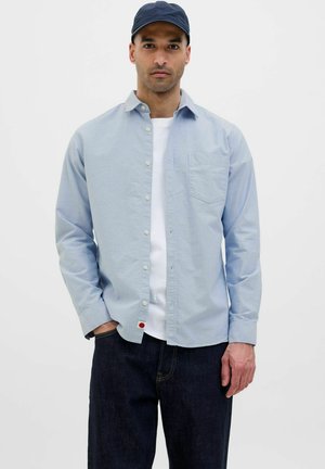 Hellblaues Button-Down-Hemd mit langen Ärmeln, Brusttasche und rotem Akzent am Saum, getragen über einem weißen T-Shirt, kombiniert mit dunklen Jeans.
