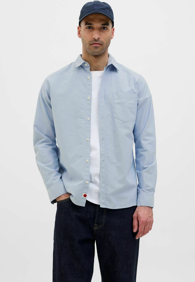 Camicia button-up azzurra con maniche lunghe, taschino sul petto e dettaglio rosso sull'orlo, indossata sopra una maglietta bianca e abbinata a jeans scuri.