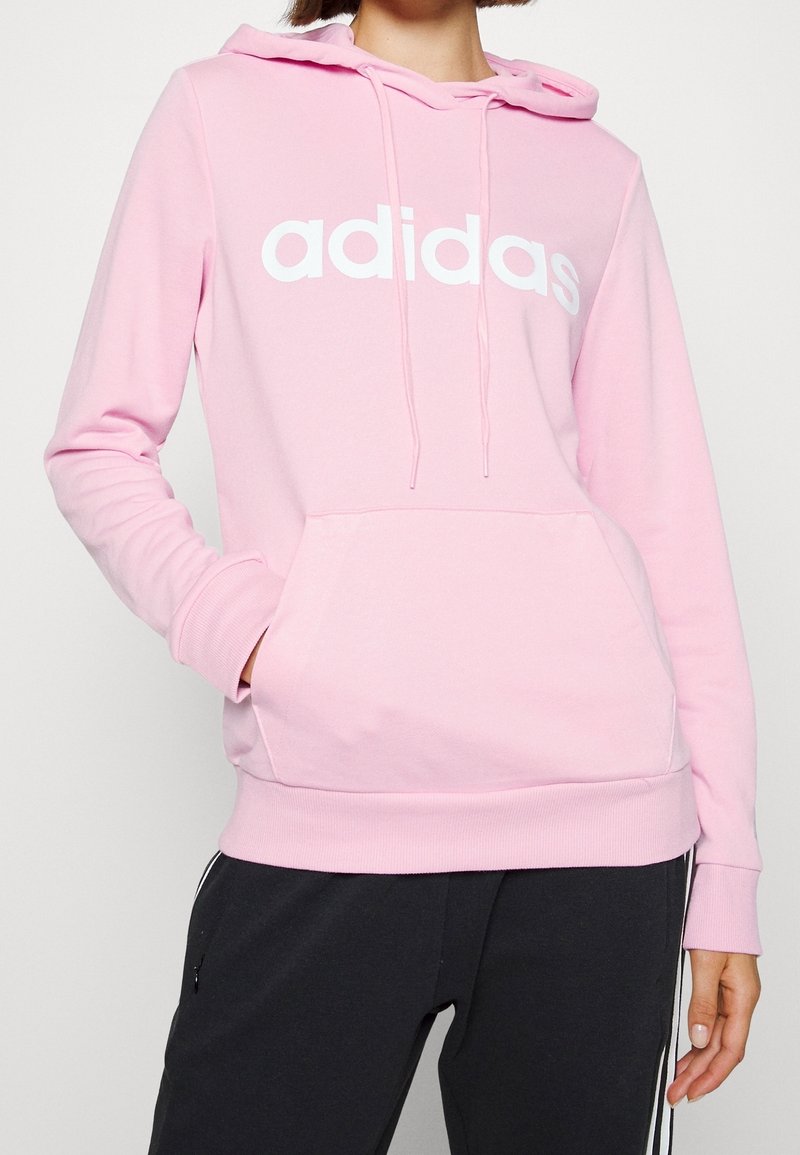 Jasnoróżowa bluza z kapturem z przednią kieszenią, białym logo adidas, regulowanym kapturem ściąganym sznurkiem oraz ribbami przy mankietach i dolnym brzegu. Miękka faktura materiału.