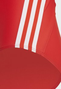 Bañador rojo con diseño de tres rayas blancas. La tela parece suave y elástica, con un corte de cintura alta. Los detalles incluyen costuras cosidas.