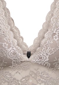 Ελαφριά μπεζ σουτιέν από δαντέλα με λουλουδένια σχέδια, κυματιστές άκρες και μια μικρή μαύρη ετικέτα μάρκας στο κέντρο μπροστά.