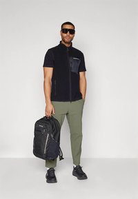 Vyras su saulės akiniais, apsivilkęs juodą liemenę ir žalias kelnes, laiko juodą "Columbia" kuprinę priešais vientisą foną.