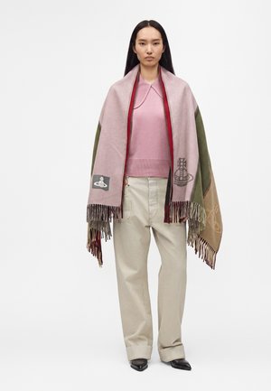 Femme portant un pull rose et un pantalon beige, avec un grand châle multicolore à franges sur les épaules.