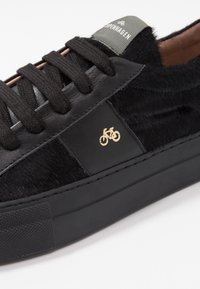 Svarta sneakers som har läder och fuskpäls, runda skosnören, en texturerad gummisula och en gyllene cykelsymbol på sidan.