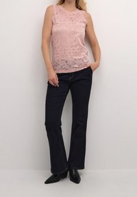 Růžový krajkový top bez rukávů s květinovým vzorem, kombinovaný s tmavě modrými rozšířenými džíny. Modelka má na sobě černé kotníkové boty na podpatku. Měkká textura a přiléhavý střih.