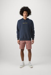 Felpa navy con testo giallo, tasca a marsupio e cappuccio con cordino, abbinata a pantaloni corti rosa chiaro e sneakers bianche su uno sfondo neutro.