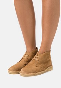 Botas de tobillo de ante en color marrón claro con punta redondeada, dos ojales para los cordones y una suela de goma en un tono marrón claro contrastante. Acabado de superficie texturizada.