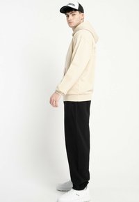 Młody mężczyzna w beżowej bluzie z kapturem, czarnych spodniach, białych sneakersach i czarno-białej czapce stoi bokiem na jednolitym białym tle.
