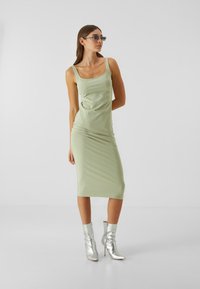 Robe midi sans manches vert menthe, avec une coupe ajustée et une texture plissée. Associée à des bottines argentées brillantes. Lunettes de soleil transparentes portées.