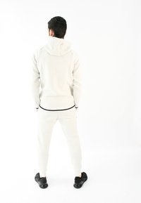 Chándal blanco con capucha; textura suave, diseño ajustado. Acentos negros en los puños y la cinturilla. Zapatillas negras completan el atuendo.