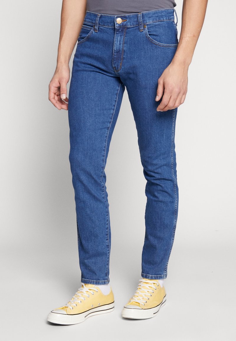 Blå denimjeans med slim fit, hög midja, fem fickor och kontrasterande sömmar. Parat med gula sneakers.