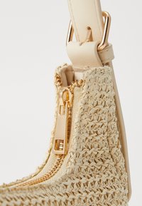 Beżowa pleciona torebka z charakterystycznym zamkiem błyskawicznym, teksturowanym materiałem przypominającym słomę oraz gładkim skórzanym paskiem z dodatkami w złotym kolorze.