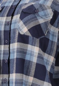 Karierter Hemd mit blauen und marineblauen Quadraten, ausgestattet mit Knöpfen, zwei Brusttaschen und einem strukturierten Stoff. Auffällige Stickdetails an den Taschen.