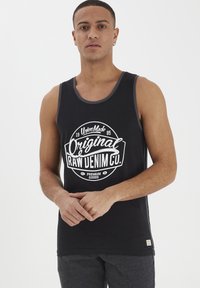 Canottiera nera con una grande grafica bianca che presenta il testo "Original Raw Denim Co.", design del logo rotondo e scollatura grigia a contrasto.