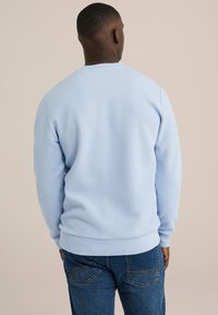 Világoskék pulóver, texturált anyagból, kerek nyakkal, bordázott mandzsettákkal és szegéllyel. Sötét farmernadrággal viselve, hátulról nézve.