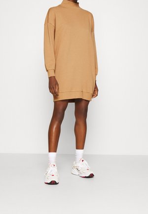 Personne portant une robe pull à manches longues couleur chameau, des chaussettes blanches et des baskets blanches avec des accents rouges, debout sur un fond uni.