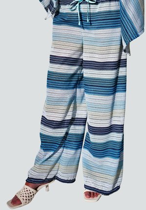 Spodnie dresowe o szerokich nogawkach z poziomymi paskami w kolorach niebieskim, białym i beżowym, zestawione z białymi sandałami na obcasie z odkrytymi palcami.