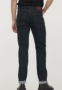 Jean en denim noir avec une coupe slim, présentant des poches arrière avec des détails de couture, des revers retournés et une étiquette en cuir à la taille.
