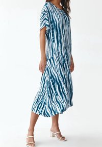 Vestido de tie-dye azul y blanco con un corte holgado, de longitud hasta la mitad de la pantorrilla, mangas cortas y textura de tela suave. Se llevan sandalias de tacón bloque.