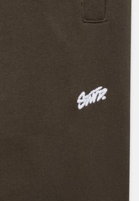 Brun bomullstyg med en slät yta, med en vit broderad logotyp i en avslappnad skrivstil. Inkluderar en liten fickdetalj.