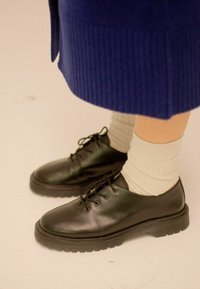 Scarpe di pelle nera con punte rotonde, design con lacci e suola in gomma spessa. Indossate con calzini a coste grigio chiaro.