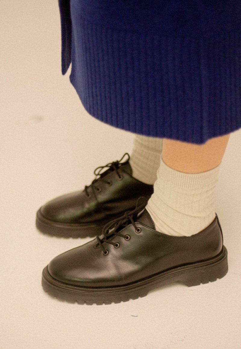 Scarpe di pelle nera con punte rotonde, design con lacci e suola in gomma spessa. Indossate con calzini a coste grigio chiaro.