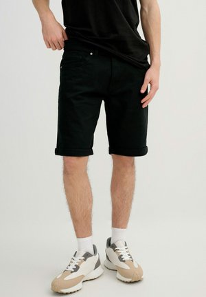 Hombre con pantalones cortos negros remangados, calcetines blancos, zapatillas beige y blancas, de pie frente a un fondo claro y liso.