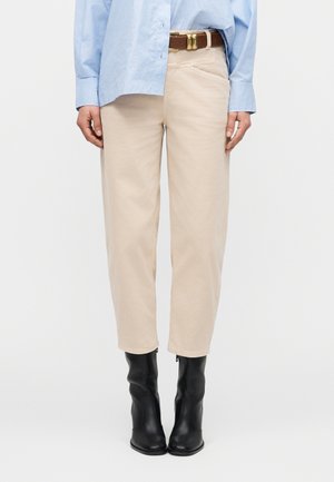 Személy bézs egyenes szárú nadrágban, barna övvel, világoskék ingben és fekete, magas sarkú bokacsizmában, sima háttér előtt.