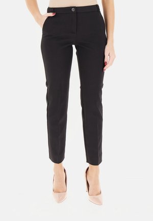 Donna che indossa pantaloni neri aderenti alla caviglia con bottone e cerniera, abbinati a tacchi alti nude con punta appuntita, mani rilassate.