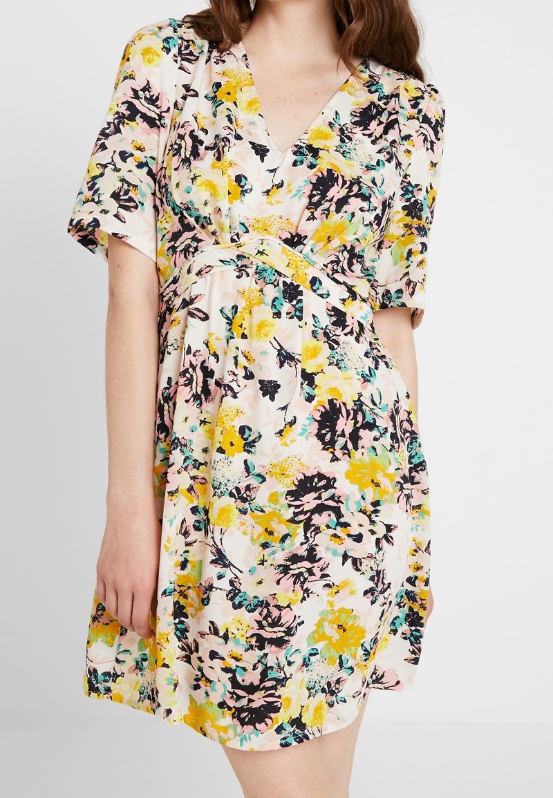 Femme portant une robe à manches courtes à fleurs jaunes, noires, roses et bleu sarcelle sur fond clair, debout avec un bras détendu.