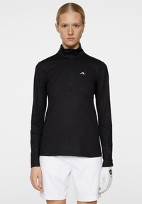 Musta pitkähihaine paita, jossa on vetoketjukaulus, ja siinä on tekstuuri kuvio sekä valkoinen logo rinnassa. Yhdistetty valkoisiin alaosiin ja hansikkaaseen.