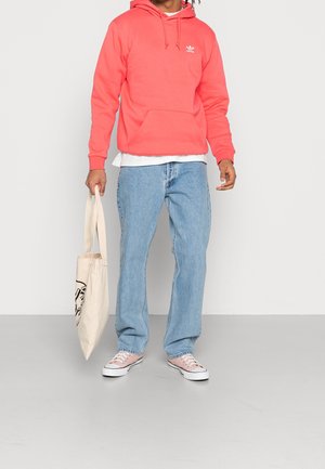 Korallfärgad hoodie med en framficka och logotyp, ihop med ljusblå jeans och vita sneakers. Håller en beige tote-väska med en grafik.