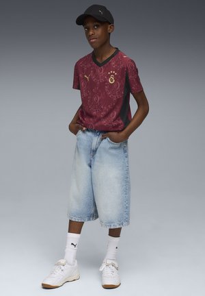 Mørkerød mønstret fodboldtrøje med sorte accenter, parret med lyseblå knælange denimshorts og hvide sneakers. En sort kasket fuldender looket.