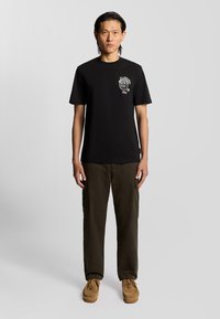 Sort bomulds t-shirt med grafisk print på venstre bryst. Parret med mørkegrønne cargo bukser og beige slip-on sko.