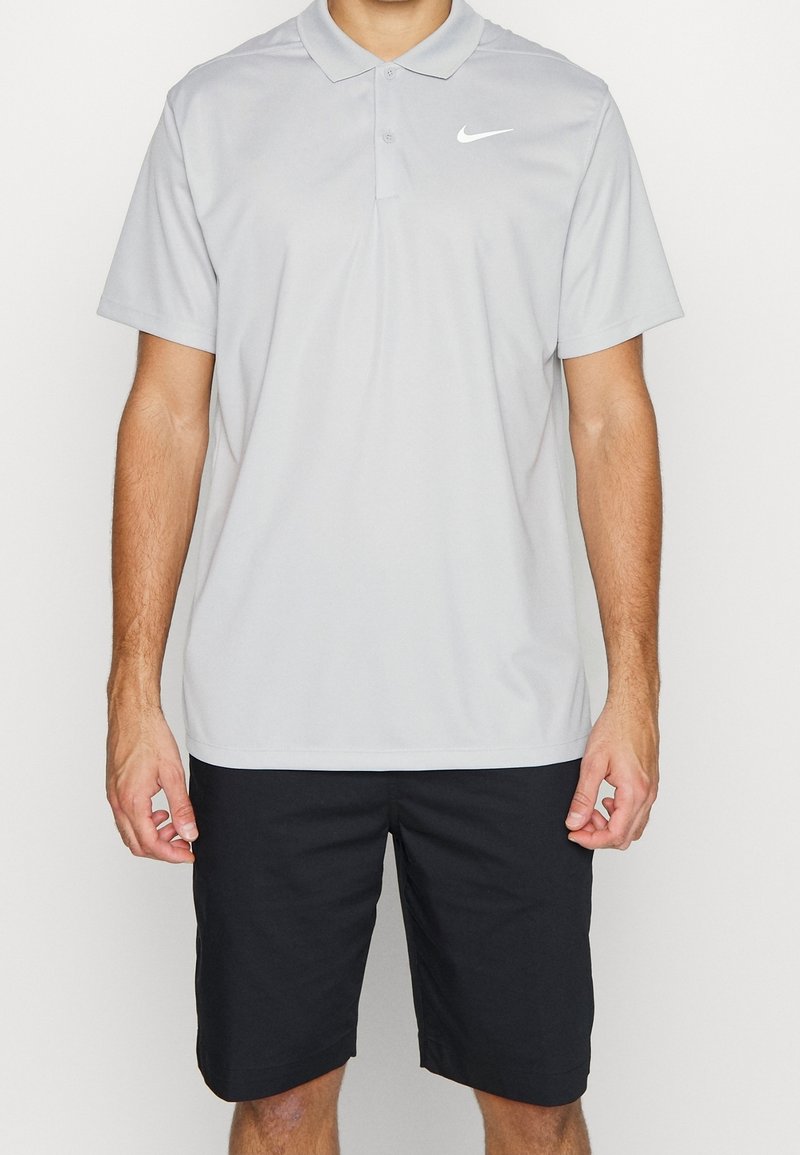 Svetle šedá polo tričko s límečkem a třemi knoflíky, s bílým logem Nike na levé části hrudi. V kombinaci s černými šortkami.