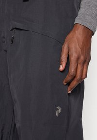 Το δεξί χέρι resting δίπλα σε σκούρες γκρι παντελόνες με φερμουάρ στην τσέπη και μικρό ανακλαστικό λογότυπο κοντά στο μηρό.