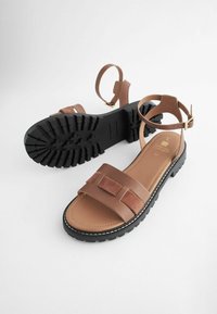 Braune Sandalen verfügen über einen breiten Vorderriemen mit Wildlederakzenten, eine gepolsterte Fußsohle und eine strukturierte schwarze Gummisohle. Ein verstellbarer Knöchelriemen ist enthalten.