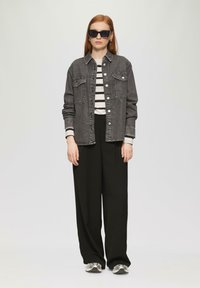Veste en denim noir avec des boutons, chemise rayée et pantalon noir à jambes larges. Le mannequin porte des lunettes de soleil surdimensionnées et des baskets en argent.