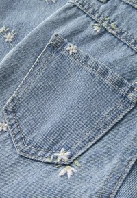 Žep iz svetlo modrega jeansa z zelenim in belim vezenim cvetličnim vzorcem, ki vsebuje natančne šive in teksturirano tkanino.