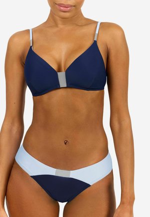 Frau trägt einen zweiteiligen Bikini in Navy und Hellblau mit dünnen Trägern und steht vor einem schlichten weißen Hintergrund.