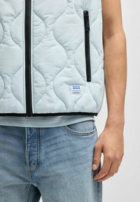 Gepolsterte hellblaue Weste mit schwarzen Reißverschlüssen und Akzenten. Verfügt über ein Markenetikett, ein strukturiertes wellenförmiges Design und ist über einem grauen Oberteil getragen.