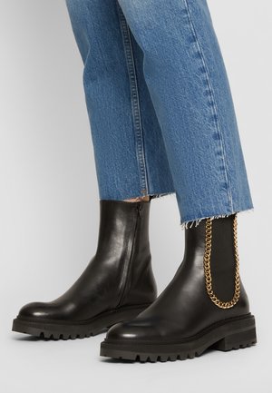 Schwarze Leder-Ankle-Boots mit einer strukturierten Sohle und goldenen Kettendetails an der Seite, kombiniert mit Jeans mit fransigem Saum.