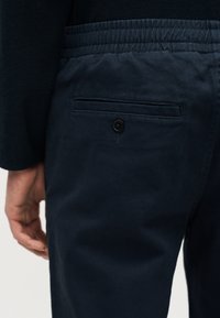 Κοντινό πλάνο σκούρου μπλε παντελονιού με ελαστική μέση και μία πίσω τσέπη με κουμπί, φορεμένο από άτομο με το χέρι χαλαρά στο πλάι.