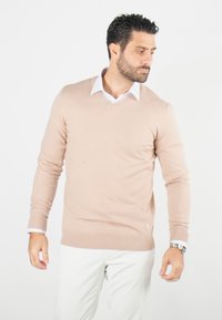 Pull en tricot beige avec un col en V, des poignets et un ourlet côtelés. Porté par-dessus une chemise blanche à col. Le modèle se tient avec une expression neutre.